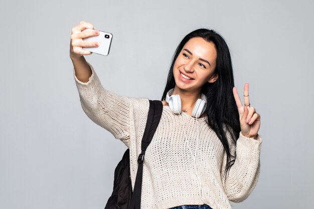 Jonge vrouw die vredesgebaar tonen terwijl het nemen van selfie op smartphone, geïsoleerd op grijze muur