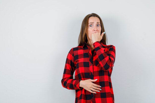 Jonge vrouw die vuist bijt, met hand op buik in geruit overhemd en angstig kijkt. vooraanzicht.