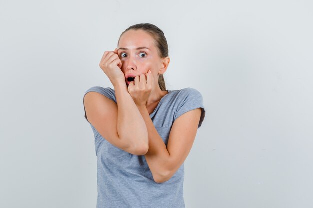 Jonge vrouw die vuisten op gezicht in grijs t-shirt houdt en bang, vooraanzicht kijkt.