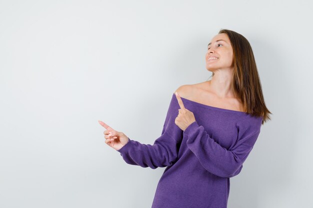 Jonge vrouw die weg in violet overhemd wijst en vreugdevol, vooraanzicht kijkt.