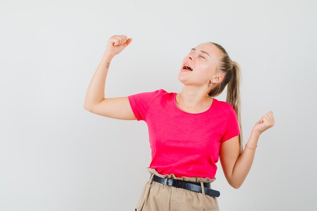 Jonge vrouw die winnaargebaar in t-shirt en broek toont en zalig kijkt