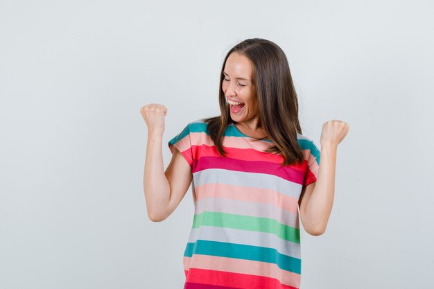 Jonge vrouw die winnaargebaar in t-shirt toont en gelukkig, vooraanzicht kijkt.