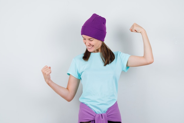 Jonge vrouw die winnaargebaar toont in blauw t-shirt, paarse muts en er gelukkig uitziet, vooraanzicht.