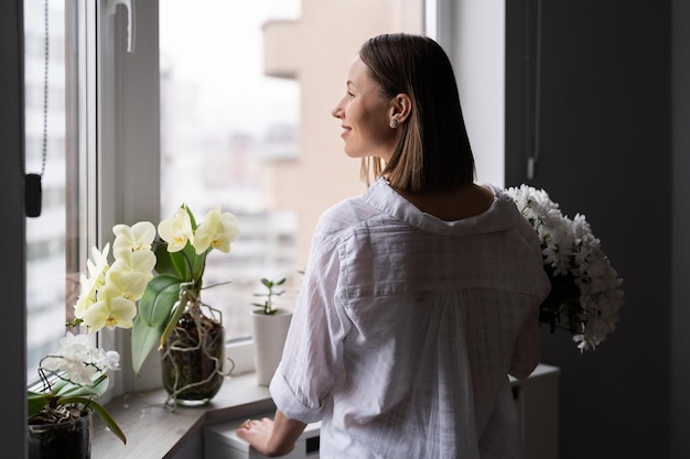 Jonge vrouw die witte vrijetijdskleding draagt en uit het raam kijkt en een boeket witte bloemen vasthoudt, wachtend op de lente of de zomer die eraan komt