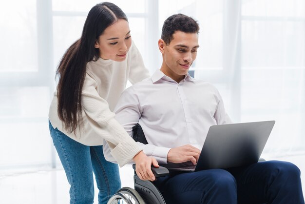 Jonge vrouw die zich achter de jonge man zit op rolstoel met behulp van laptop