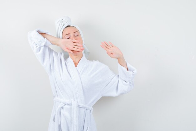 Jonge vrouw die zich bovenlichaam uitstrekt terwijl ze in witte badjas, handdoek geeuwt en ontspannen, vooraanzicht kijkt.
