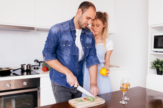 Jonge vrouw die zich dichtbij de echtgenoot bevindt die bellpepper met mes op lijst in de keuken snijdt