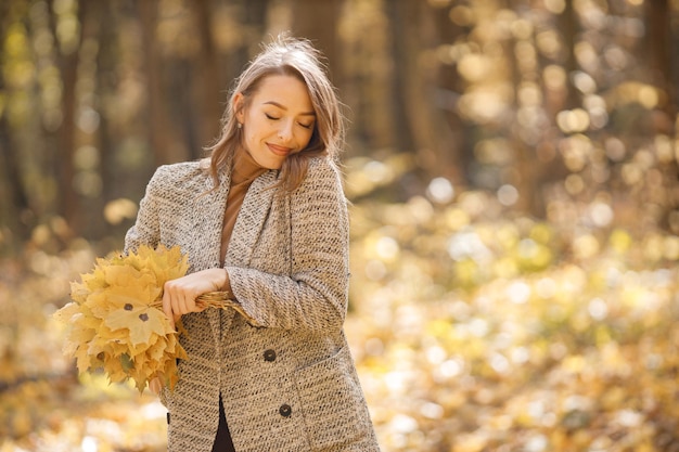 Jonge vrouw die zich in de herfstbos bevindt. Donkerbruine vrouw die gele bladeren houdt. Meisje draagt mode bruin jasje.