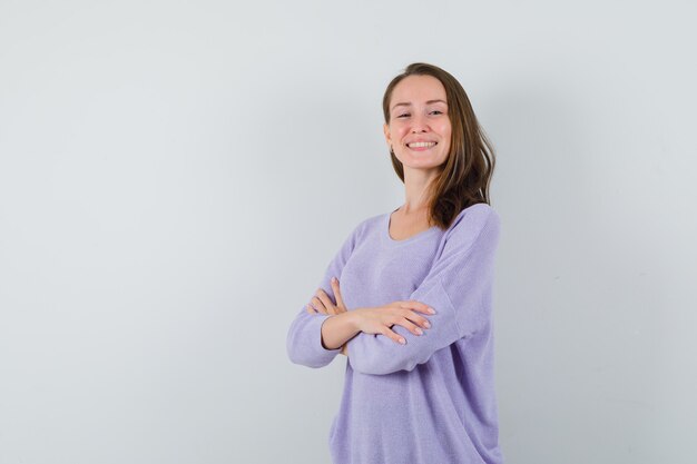 Jonge vrouw die zich met gekruiste wapens in lila blouse bevindt en tevreden kijkt. vooraanzicht.