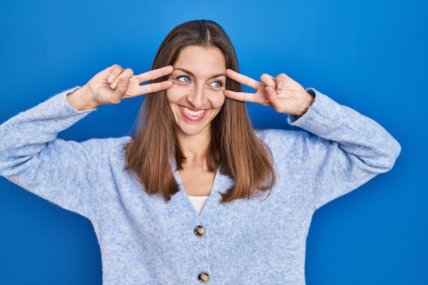 Jonge vrouw die zich over blauwe achtergrond bevindt die vredessymbool doet met vingers over gezicht dat vrolijk glimlacht en overwinning toont