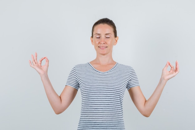 Gratis foto jonge vrouw doet meditatie met gesloten ogen in gestreept t-shirt en ziet er vredig uit. vooraanzicht.