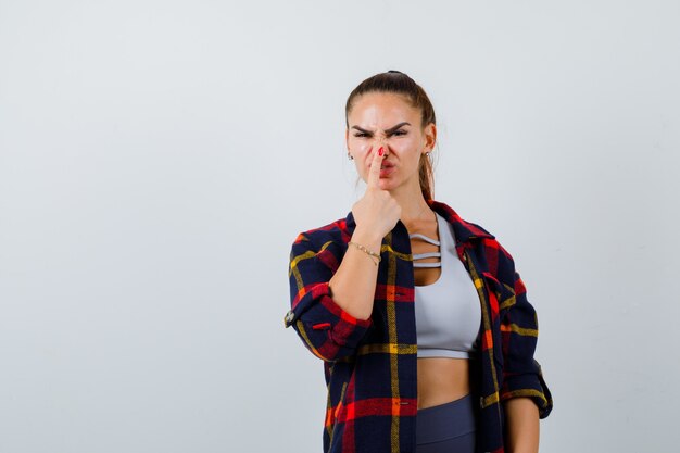 Jonge vrouw drukt vinger op neus in crop top, geruit hemd, broek en ziet er grappig uit. vooraanzicht.