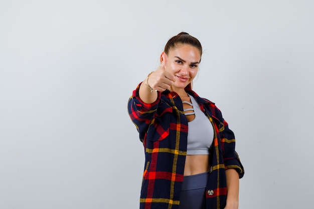 Jonge vrouw duim opdagen in crop top, geruit hemd, broek en er gelukkig uitzien. vooraanzicht.