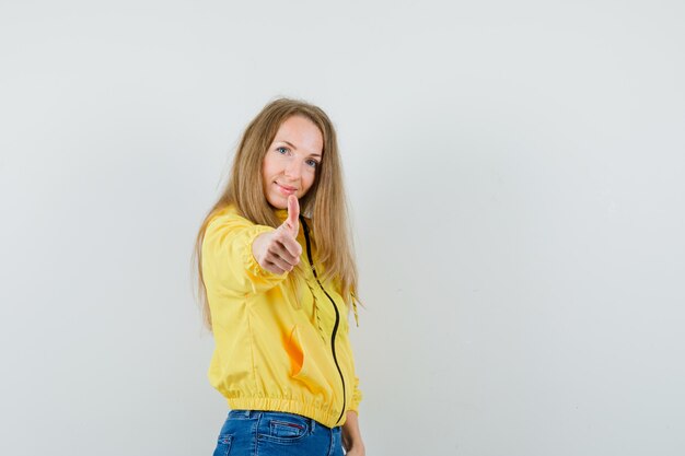 Jonge vrouw duim opdagen in gele bomberjack en blauwe jean en op zoek optimistisch. vooraanzicht.