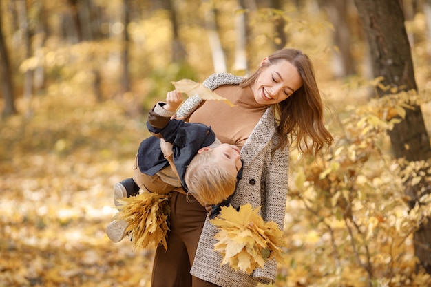 Jonge vrouw en kleine jongen in herfst bos. Vrouw die haar zoon op haar handen houdt. Jongen die modekleding draagt en gele bladeren vasthoudt.