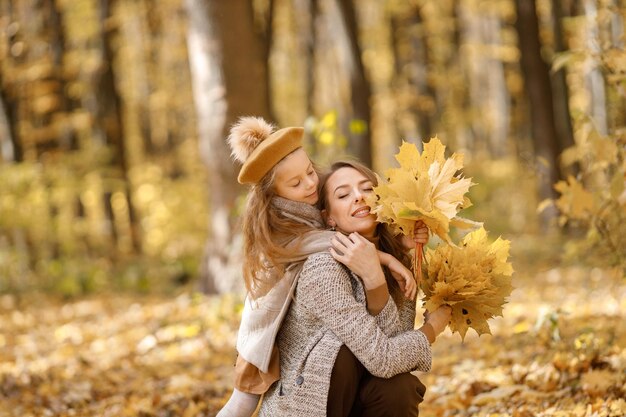 Jonge vrouw en meisje in de herfstbos. Vrouw haar dochter knuffelen. Meisje dat modekleding draagt en gele bladeren vasthoudt.