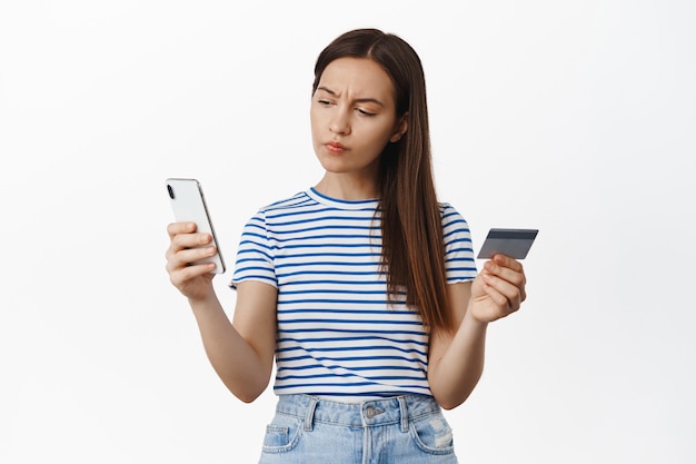 Jonge vrouw fronst, kijkt naar het smartphonescherm en houdt creditcard in de hand, denkt na, doet een doordachte aankoop, overweeg iets te kopen of niet, witte muur