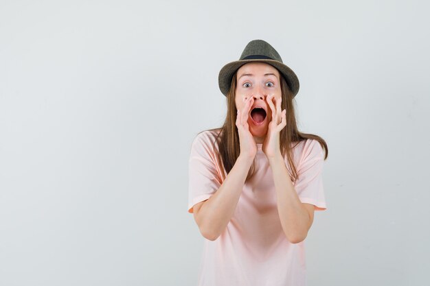 Jonge vrouw geheim vertellen met handen in de buurt van mond in roze t-shirt, hoed vooraanzicht.