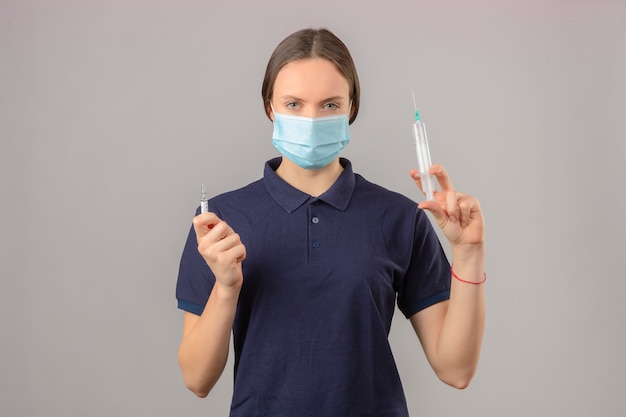 Jonge vrouw, gekleed in blauw poloshirt in beschermende medische masker met spuit en medicijn vaccin fles kijken camera met ernstige gezicht staande op geïsoleerde grijze achtergrond