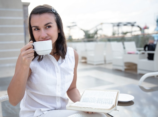 Jonge vrouw genieten van koffie in de ochtend op een open zomerterras met een boek in haar handen