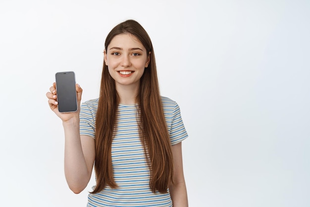 Jonge vrouw glimlachend gelukkig, smartphone leeg scherm, applicatie-interface tonen. Concept van mobiele winkel, online bankieren of winkelen, witte achtergrond.