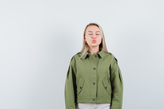 Jonge vrouw haar lippen pruilen tijdens het sluiten van de ogen in groene jas, spijkerbroek, vooraanzicht.