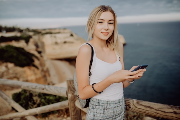 Jonge vrouw haar sociale netwerkstatus bijwerken terwijl u ontspant op het rotsstrand met panoramisch uitzicht