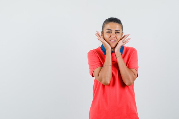 Jonge vrouw hand in hand op de wang in rood t-shirt en ziet er mooi uit, vooraanzicht.