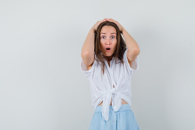 Jonge vrouw hand in hand op het hoofd in witte blouse en lichtblauwe rok en verbaasd kijken