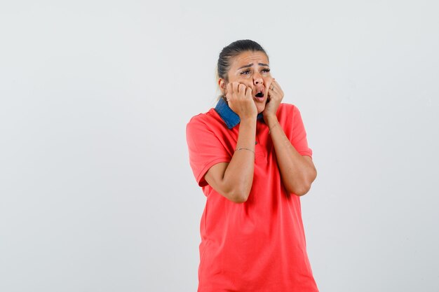 Jonge vrouw hand in hand op wangen in rood t-shirt en op zoek verrast. vooraanzicht.