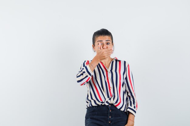 Jonge vrouw hand op mond zetten in gestreepte blouse en verbaasd kijken.