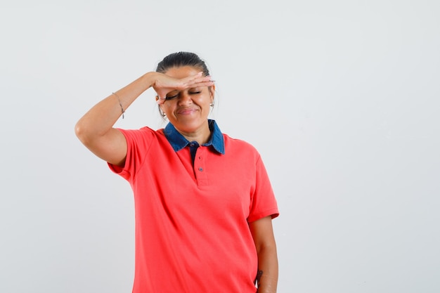 Jonge vrouw hand op voorhoofd in rood t-shirt zetten en gekwelde op zoek. vooraanzicht.