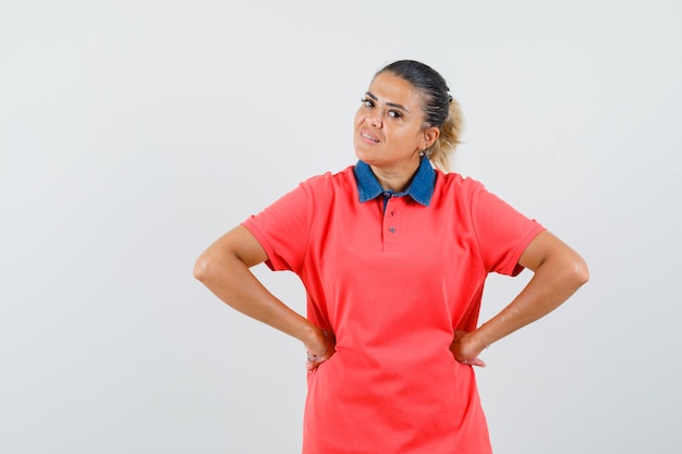 Jonge vrouw handen op taille in rood t-shirt zetten en mooi, vooraanzicht kijken.