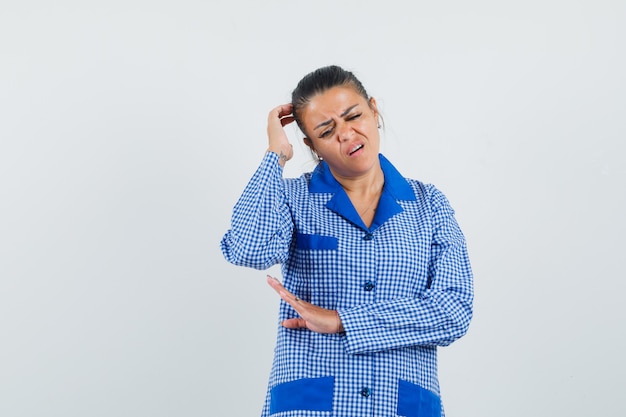 Jonge vrouw hoofd krabben terwijl staande in denken pose in blauw pastel pyjama shirt en peinzend kijken. vooraanzicht.