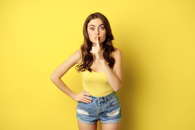 Gratis foto jonge vrouw houdt geheim, toont stilte, stil gebaar, sussend, vinger op lippen, staande over gele achtergrond