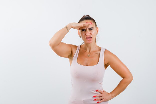 Jonge vrouw houdt hand op het hoofd op zoek naar manier terwijl ze hand op de heup in witte tanktop houdt en verbaasd kijkt. vooraanzicht.