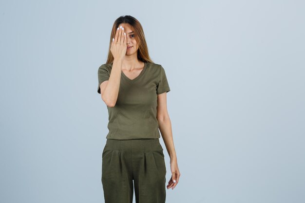 Jonge vrouw houdt hand op oog in t-shirt, broek en ziet er optimistisch uit. vooraanzicht.