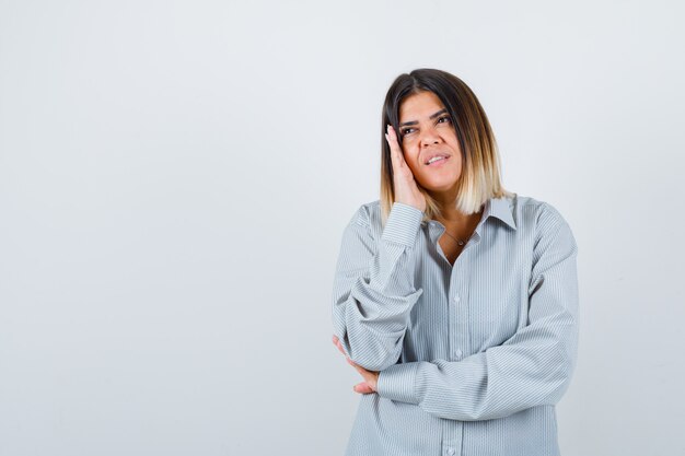 Jonge vrouw houdt hand op wang in oversized shirt en ziet er mooi uit, vooraanzicht.