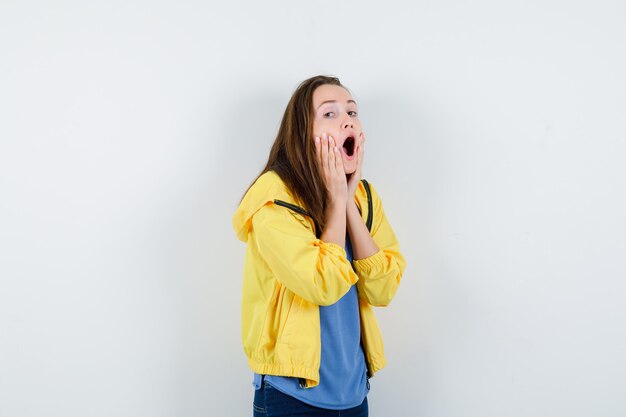 Jonge vrouw houdt handen op wangen in t-shirt, jas en kijkt verrast. vooraanzicht.