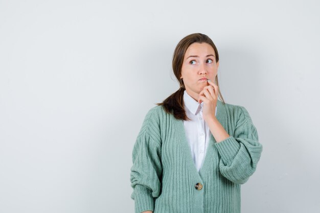 Jonge vrouw houdt vinger in de buurt van mond, kijkt omhoog in blouse, vest en kijkt peinzend, vooraanzicht.