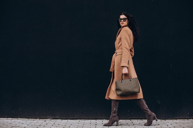 Jonge vrouw in beige jas die buiten op zwarte muurachtergrond loopt walking