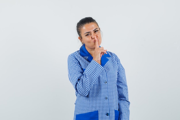 Jonge vrouw in blauw pastel pyjama shirt wijsvinger op lip zetten, stilte gebaar tonen en mooi, vooraanzicht kijken.