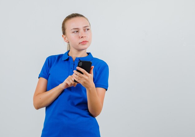 Jonge vrouw in blauw t-shirt met behulp van mobiele telefoon en peinzend op zoek