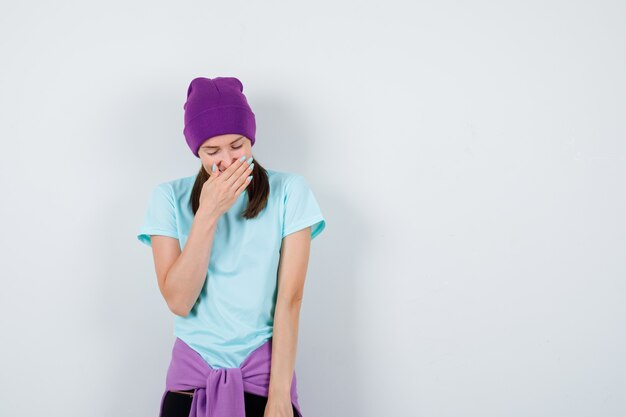 Jonge vrouw in blauw t-shirt, paarse muts die mond bedekt met hand, lachend en vrolijk kijkend, vooraanzicht.