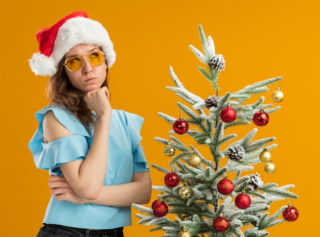 jonge vrouw in blauwe boven- en kerstmuts met gele bril opzoeken verbaasd staande naast een kerstboom over oranje achtergrond