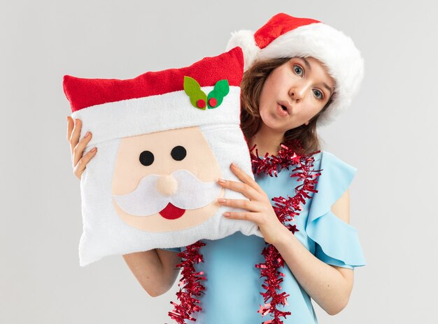 Jonge vrouw in blauwe bovenkant en santahoed met klatergoud om haar hals die het hoofdkussen van Kerstmis houden verbaasd kijken