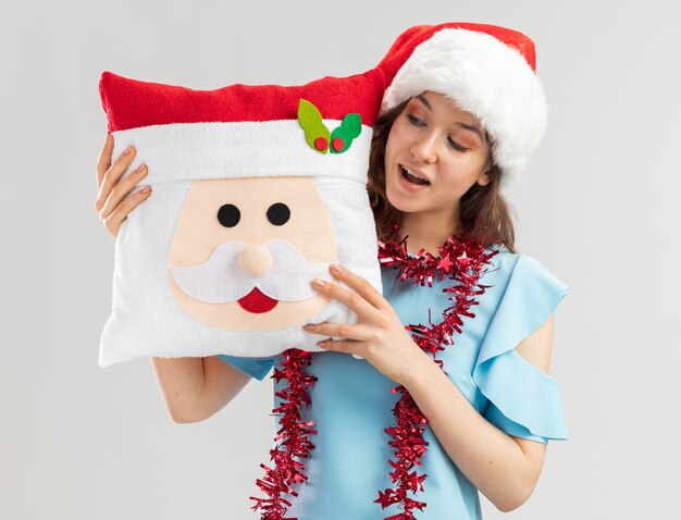 Jonge vrouw in blauwe bovenkant en santahoed met klatergoud om haar hals die het Kerstmishoofdkussen met glimlach op gezicht bekijkt