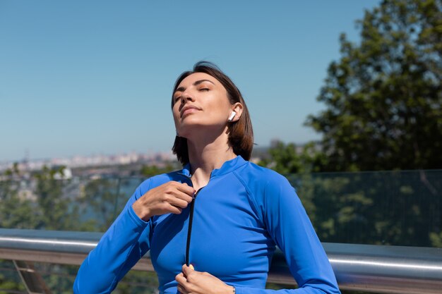 Jonge vrouw in blauwe sportkleding op brug op warme zonnige ochtend met draadloze koptelefoon ritst jas open