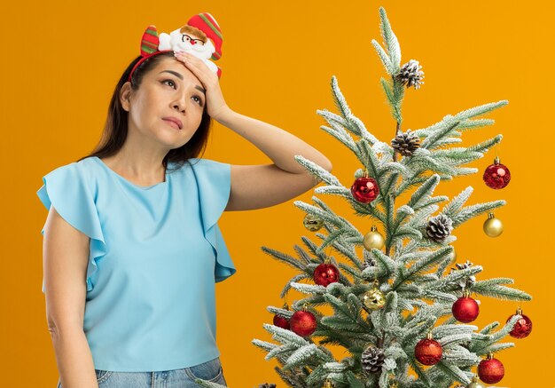 Jonge vrouw in blauwe top die grappige kerstrand op het hoofd draagt en kijkt omhoog verward met de hand op haar hoofd staande naast een kerstboom over oranje achtergrond