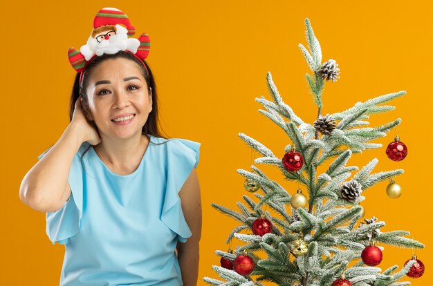 Jonge vrouw in blauwe top dragen grappige kerst rand op hoofd kijken camera lachend met blij gezicht staande naast een kerstboom over oranje achtergrond